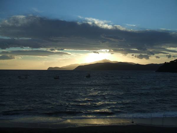 Sun setting behind dark mountains over the sea, small boats on rippled water