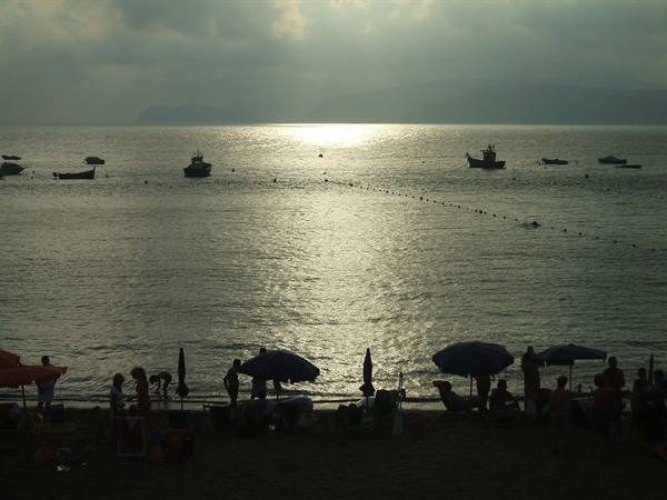 Boats anchored offshore above a crowded beach with umbrellas under hazy sun
