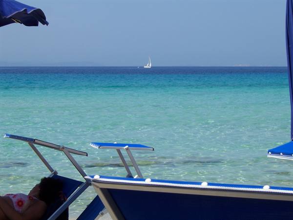 Blue umbrellas and loungers by turquoise sea, a sailboat on the horizon