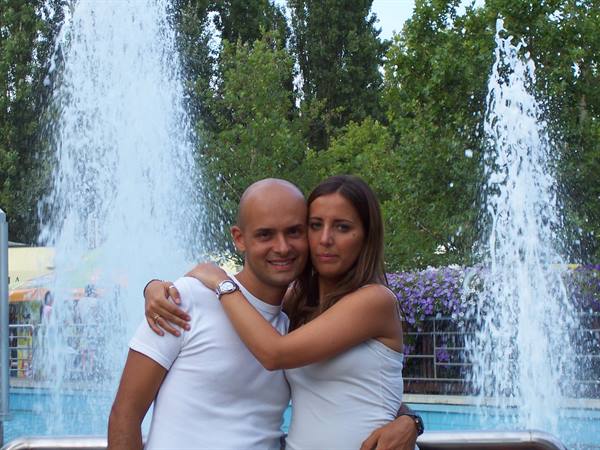 Couple in white embracing in front of a fountain in a tree-lined park