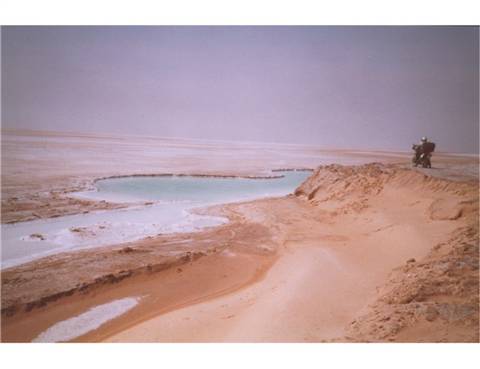Turquoise spring amid sand and salt crust, a man riding a motorcycle on the dune