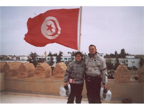 Two motorcyclists holding helmets beneath the Tunisian flag, brick ramparts behind