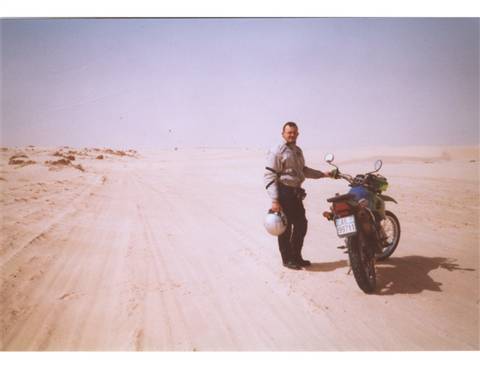 Man holding helmet beside a motorcycle on a sandy desert track