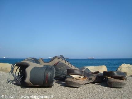 Hiking shoes and sandals on grey stone, blue sea with ships in the distance