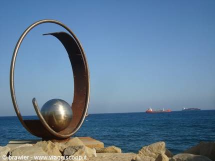 Metal sculpture with shiny sphere on rocky shore, cargo ships at sea