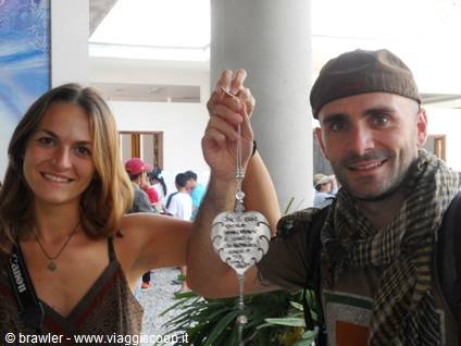 He holds up a white decorated heart ornament, she smiles, in a columned hall
