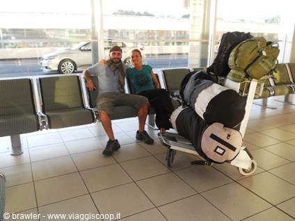 Two smiling travelers beside a luggage cart with backpacks and bags, in an airport