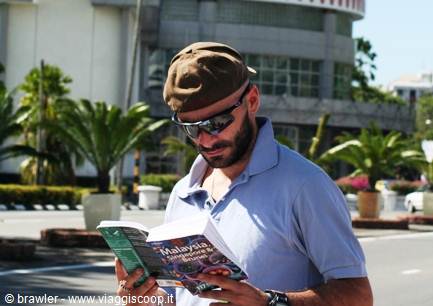 Man in brown cap and sunglasses reads a Malaysia guidebook among palm trees