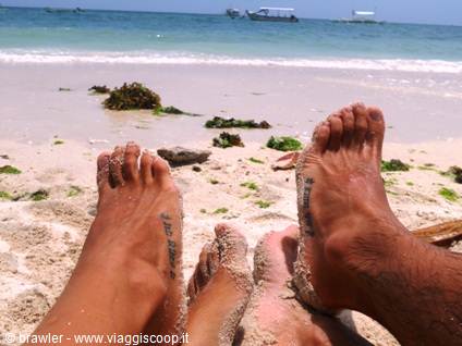 Bare tattooed feet on white sand, boats offshore on turquoise sea