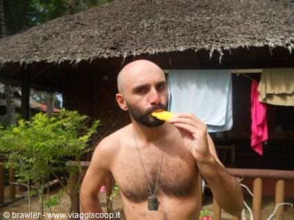 Shirtless man eating pineapple in front of thatched hut with hanging laundry