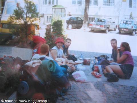 Group of backpackers eating on the sidewalk beside backpacks and sleeping mats