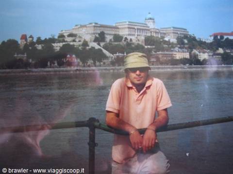 Man in bucket hat and pink polo leaning on a railing before Buda Castle on the Danube