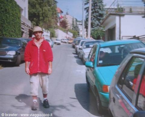 Man in red jacket and hat walks past parked cars on a sloped street