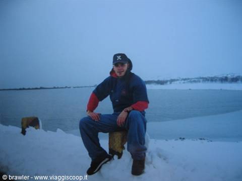 Young man in cap sitting on a wooden post in the snow by a frozen fjord