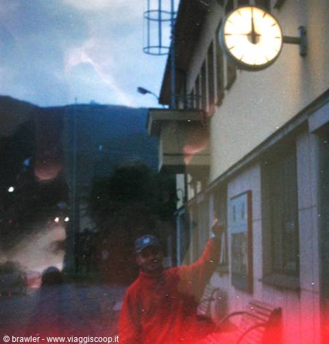 young man in red jacket points at a large illuminated clock on the station facade