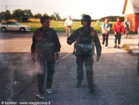 Two skydivers in jumpsuits and helmet walk on asphalt, others follow