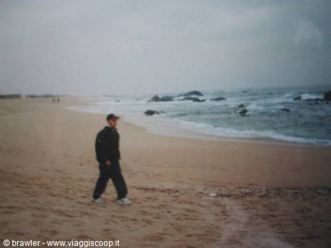 Man in dark jacket walking on grey sand beach, waves and rocks