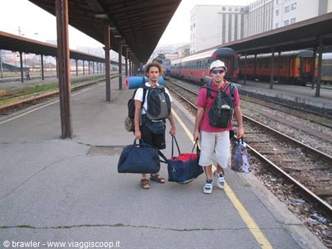 Two young backpackers with duffel bags on the station platform beside a red train