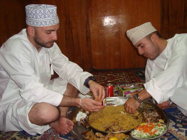 Two Omani men in white dishdasha and embroidered caps eat rice by hand
