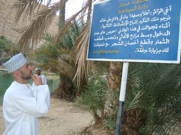 Man in white dishdasha reading blue Arabic sign among palm trees, Wadi Shab