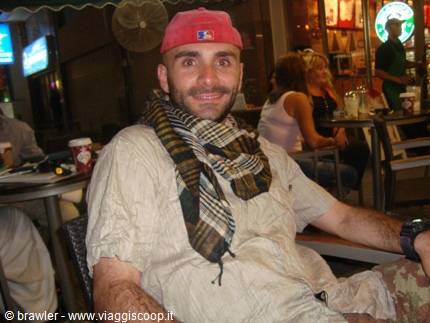 Smiling man wearing red cap and checkered keffiyeh scarf sits at evening bar