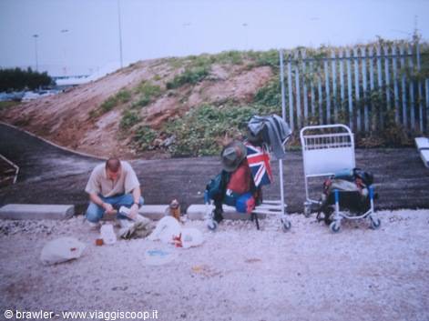 Man crouching packs luggage beside carts and a Union Jack flag at a rest stop