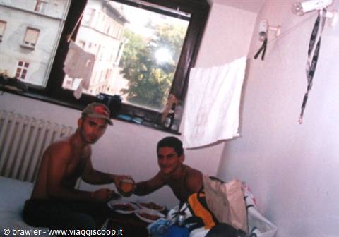 Two shirtless young men eating on a bed, towels hanging from the window