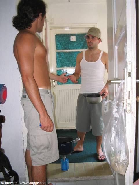 Two guys in tank tops cooking on a portable gas stove in a hostel hallway