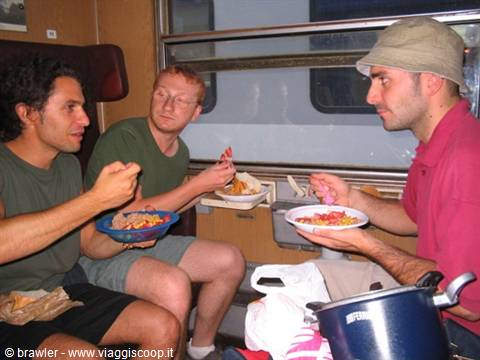 Three young men eating pasta from paper plates inside a train compartment