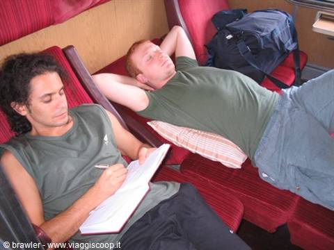 Two young men on red train seats, one writing in a notebook, one sleeping