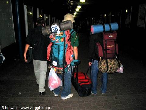 Three backpackers with sleeping mats walk at night under station lights