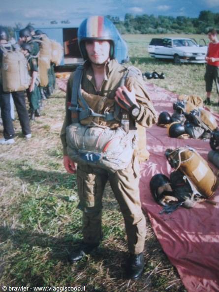 Parachutist wearing helmet and harness on grassy field, gear laid on red tarps