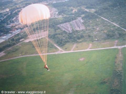 Parachutist under cream canopy descending onto green field near hangars