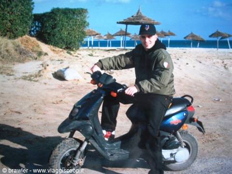 Man in green jacket on black scooter, sandy beach with straw umbrellas behind