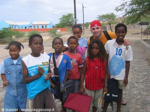 Traveler with red backpack posing with smiling local children on cobblestone street, blue building behind
