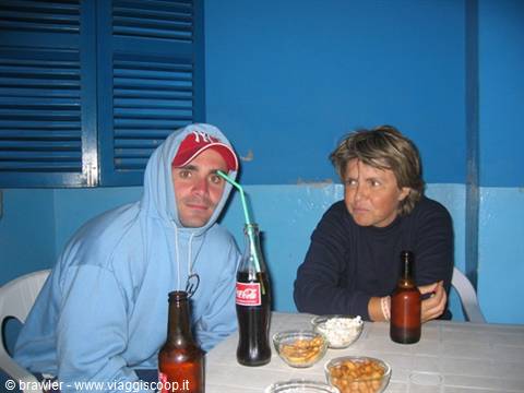 Man in hoodie and woman at table with Coke bottle, beers and snacks, blue walls