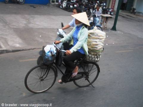 Woman in conical straw hat cycling with loaded bicycle on busy street