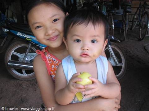 Smiling girl hugging a baby holding an apple, motorbikes parked behind