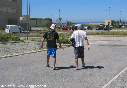 Two young men walk in flip-flops on an asphalt road toward the sea