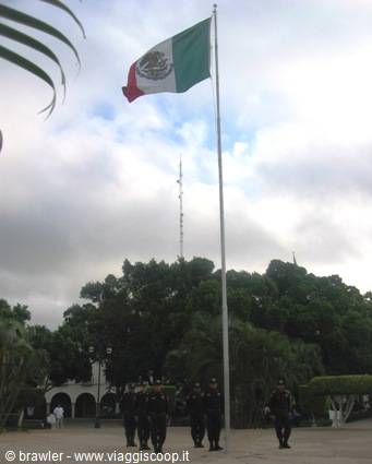 Soldiers lower the Mexican flag from a tall flagpole in a palm-lined plaza