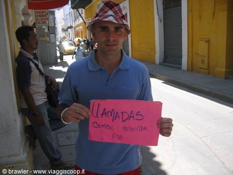 Tourist in bucket hat and blue polo holds pink sign reading Llamadas on narrow street