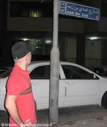 Tourist in red shirt reads the bilingual Addi Bin Zaid street sign at night