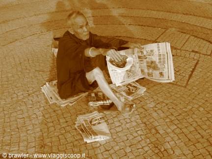 Man sitting on cobblestones reads a newspaper wrapped in dark cloths