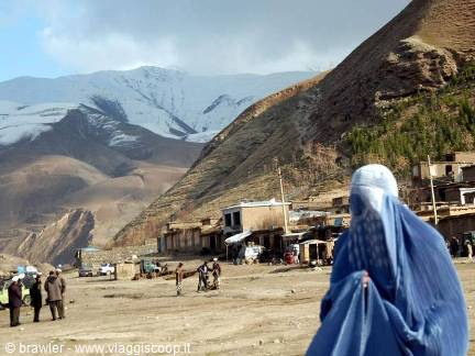 Woman in blue burqa walks along the dirt road of a village beneath snowy mountains