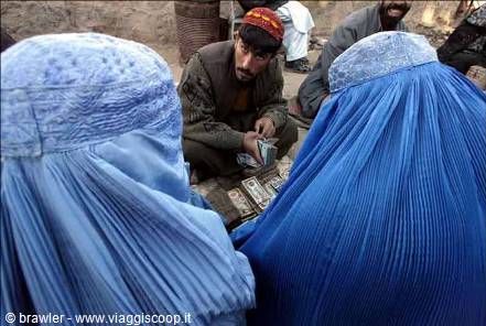Women in blue burqas buy banknotes from a vendor at a dusty market