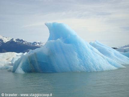 Blue iceberg floating on glacial water with snow-capped mountains behind