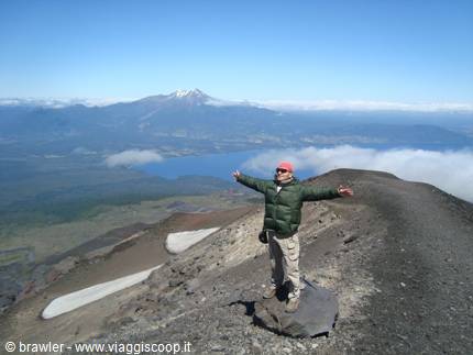 Hiker in green jacket arms outstretched on volcanic ridge, lake and snowy volcano behind