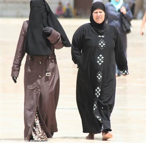 Two women walk barefoot across a light stone plaza, one in black niqab