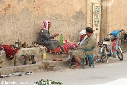 Men in keffiyeh chat beside traditional braziers and furniture on a dusty street