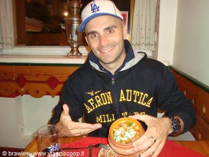 Man in a cap smiles at a wooden restaurant table, showing a bowl of soup
